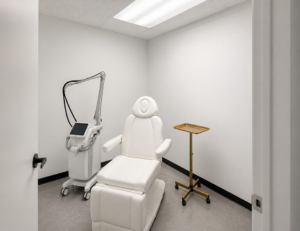 An empty treatment room with a white reclining chair, a brass tray stand, and a laser device in the corner. A laser for pigment sends short pulses of light into the spot, and the body clears the broken-up pigment over the following weeks.